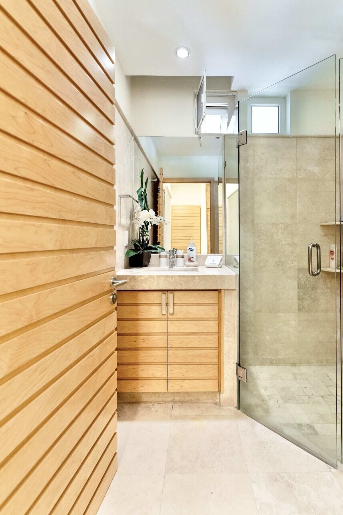 A modern bathroom with wooden cabinetry, a glass shower enclosure, and a countertop with toiletries.
