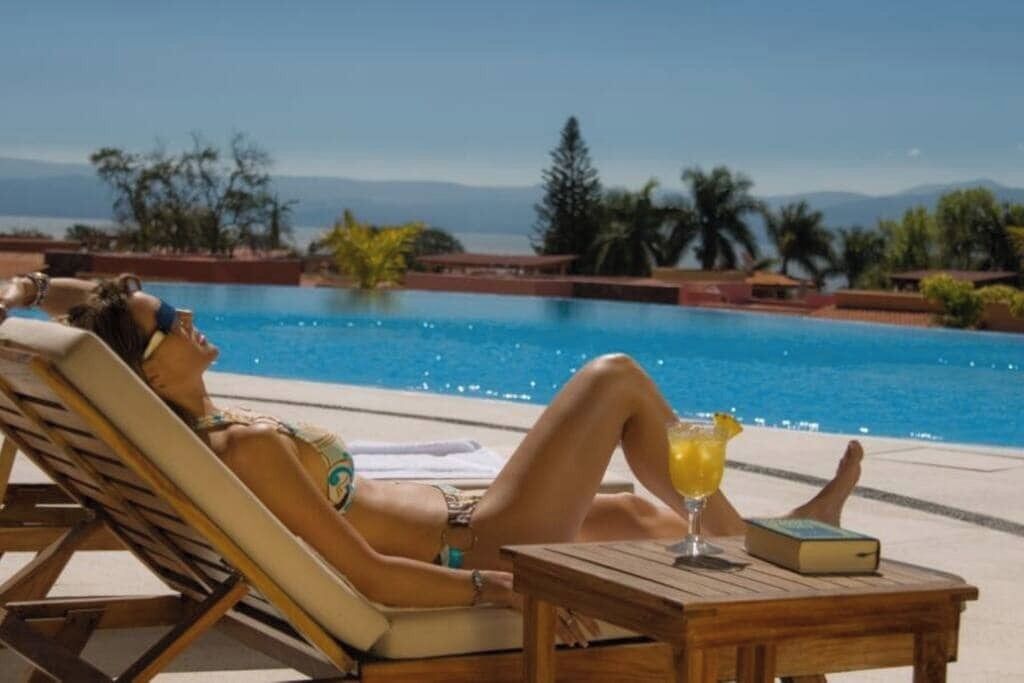 A person relaxing by a pool with a drink and a book, surrounded by palm trees and a clear view of the mountains.