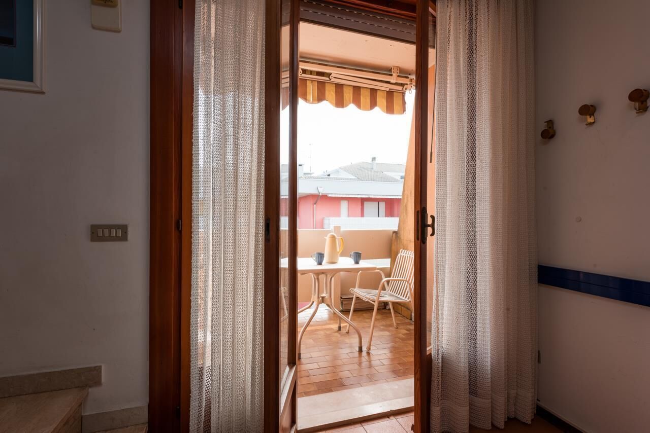 Ein Durchgang zu einem Außenbalkon mit Tisch und Stühlen, ein Fenster mit gestreiftem Sonnenschirm und einer Wandleuchte.