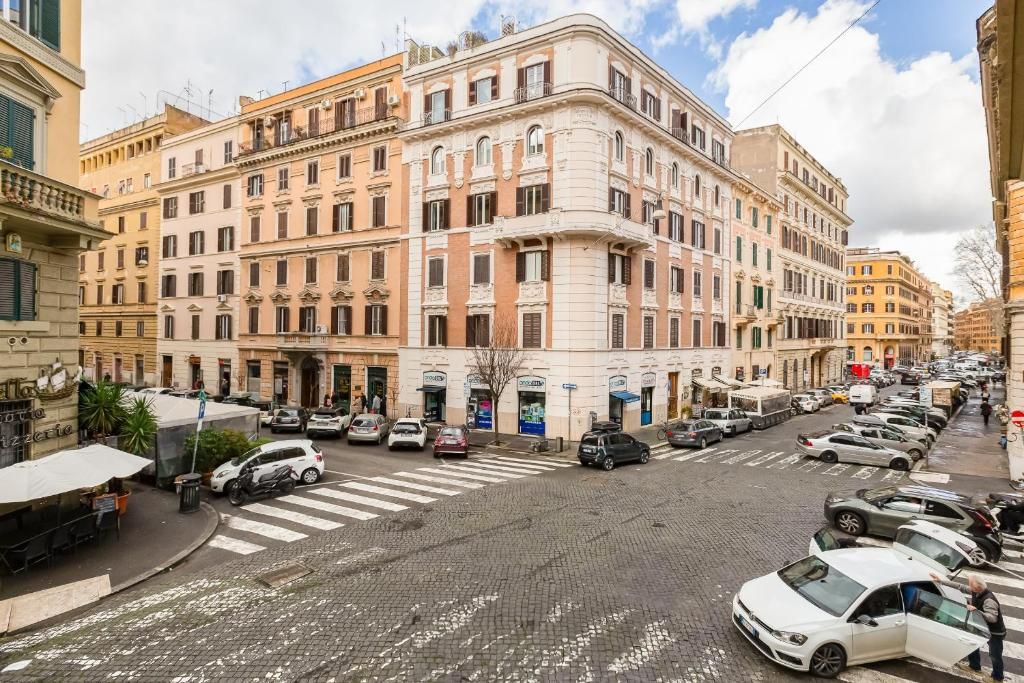 A street view with a row of parked cars, a pedestrian crossing, and buildings with classical architecture.
