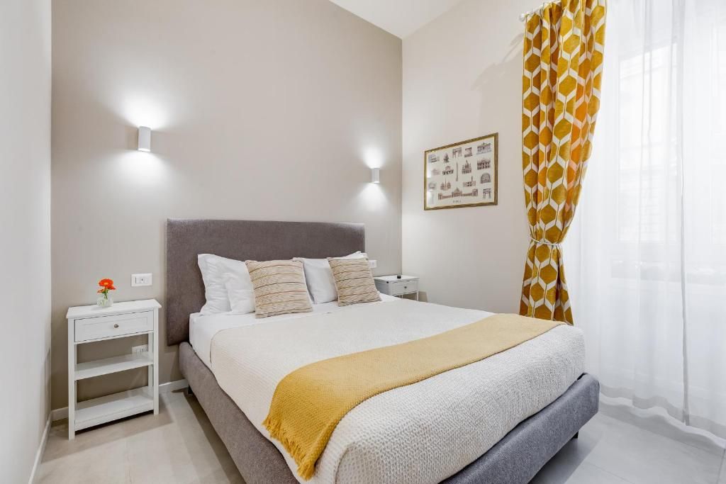 A bedroom with a bed, bedside tables, a nightstand, a wall-mounted light, and a window with curtains.