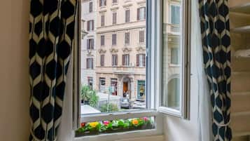 A window with patterned curtains, a view of a building with multiple stories and balconies, and a small balcony with flowers.