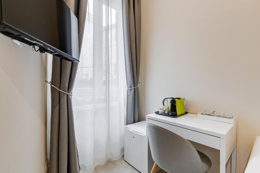 A small, modern room with a white desk, a chair, a television mounted on the wall, and a window with grey curtains.