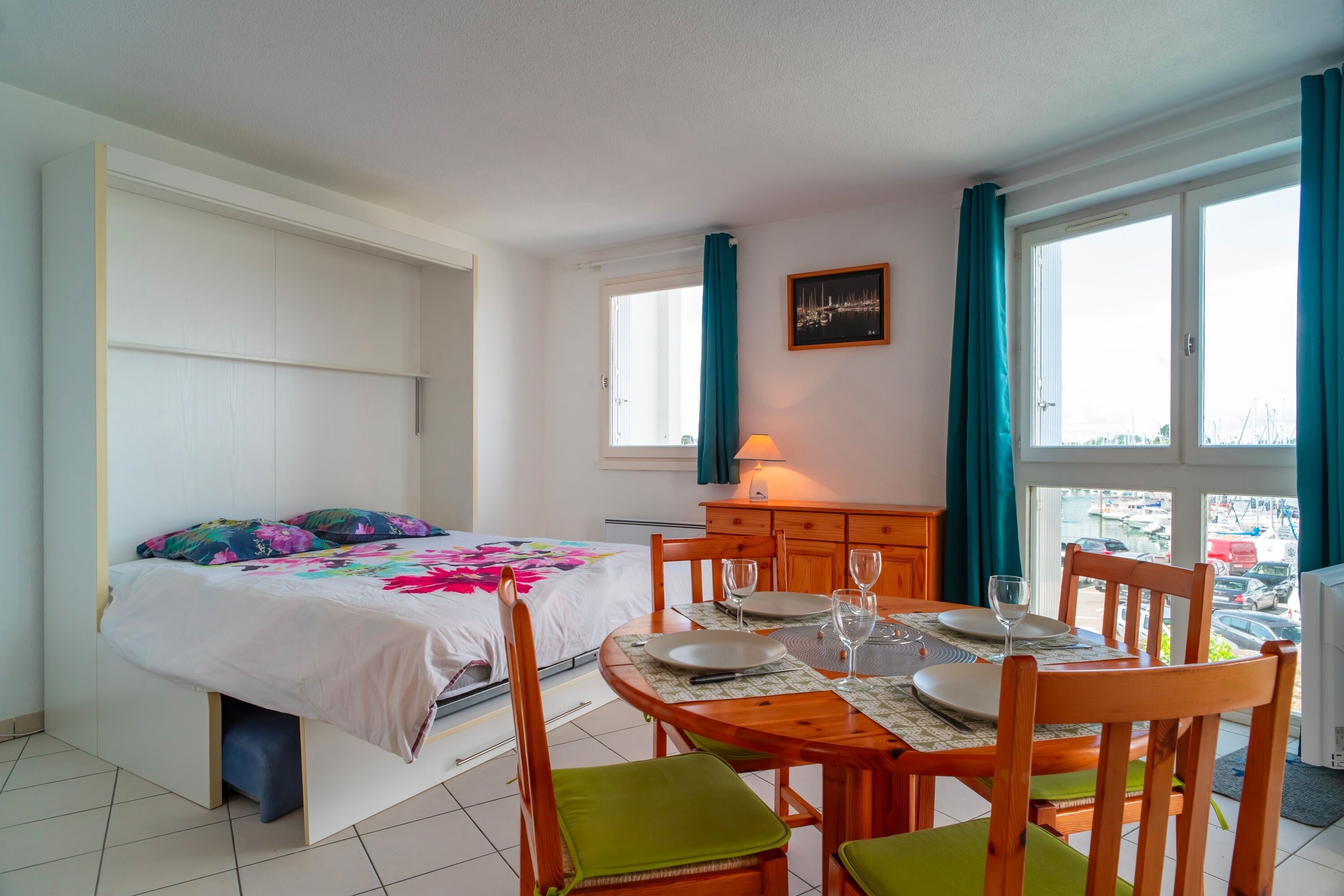A bedroom with a bed, a dining table with chairs, a window with curtains, and a view of the outside.