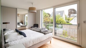 A modern bedroom with a large bed, a bedside table, a lamp, and a view of the outside. - Villa Ker Jade - vue sur la mer (Lancieux)