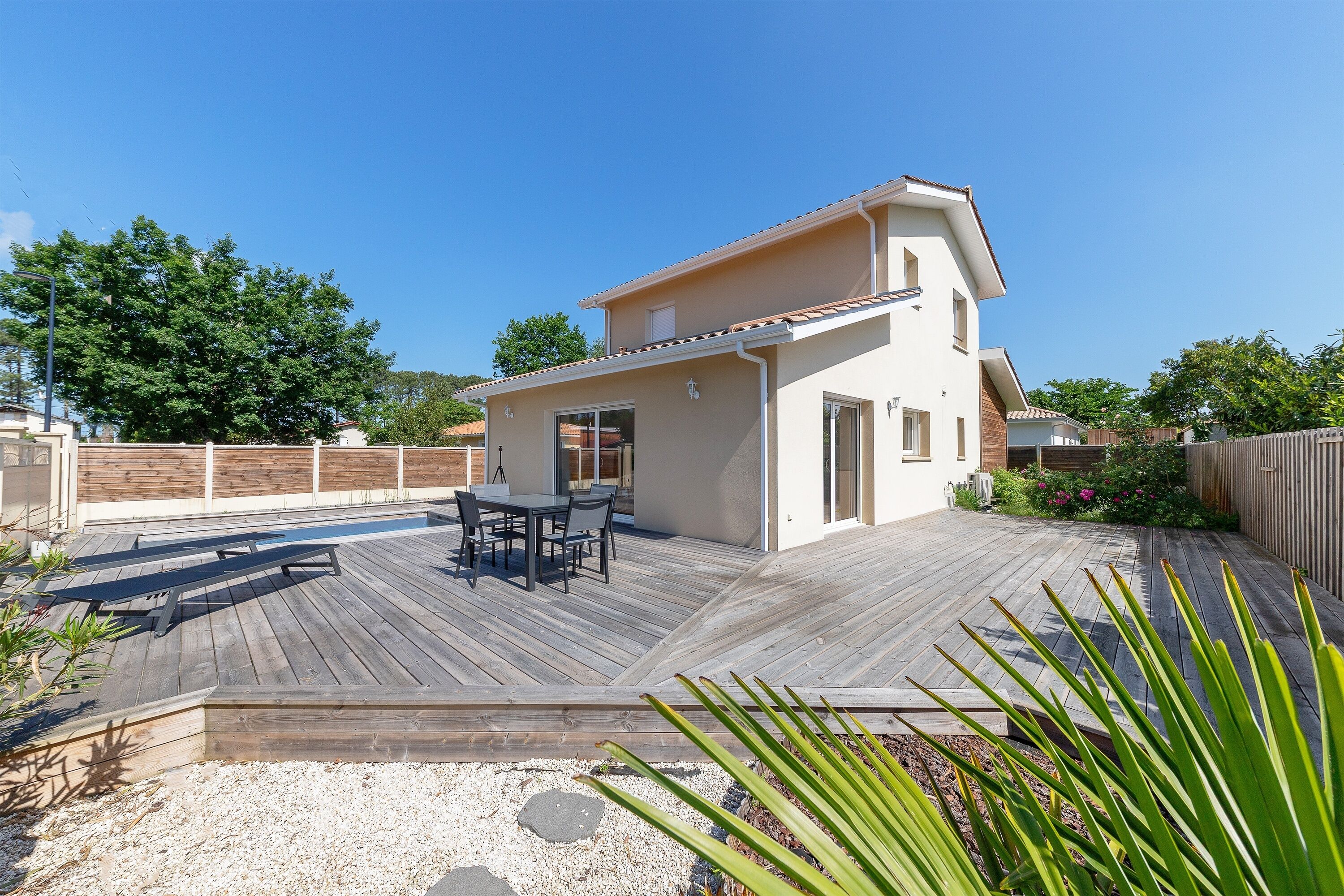 Una moderna casa a due piani con una veranda in legno, un patio con mobili da esterno e un piccolo giardino.