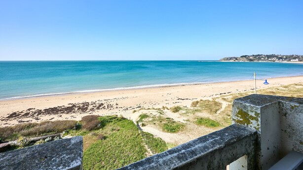 A beach with clear blue water, sandy shore, and a distant hill with buildings.