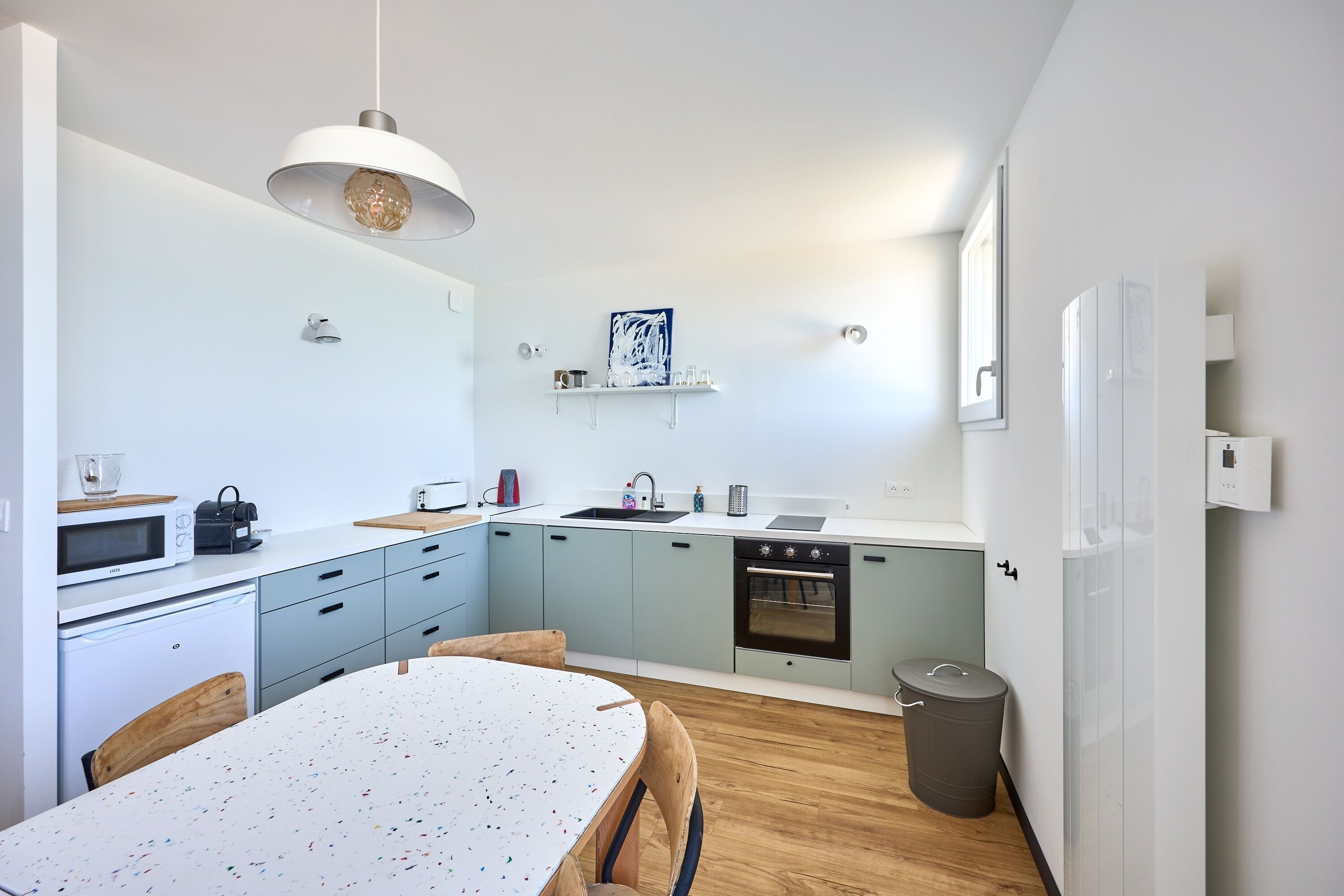 Une cuisine moderne avec des meubles bleu clair, un plan de travail blanc, une table à manger en bois et un luminaire suspendu.