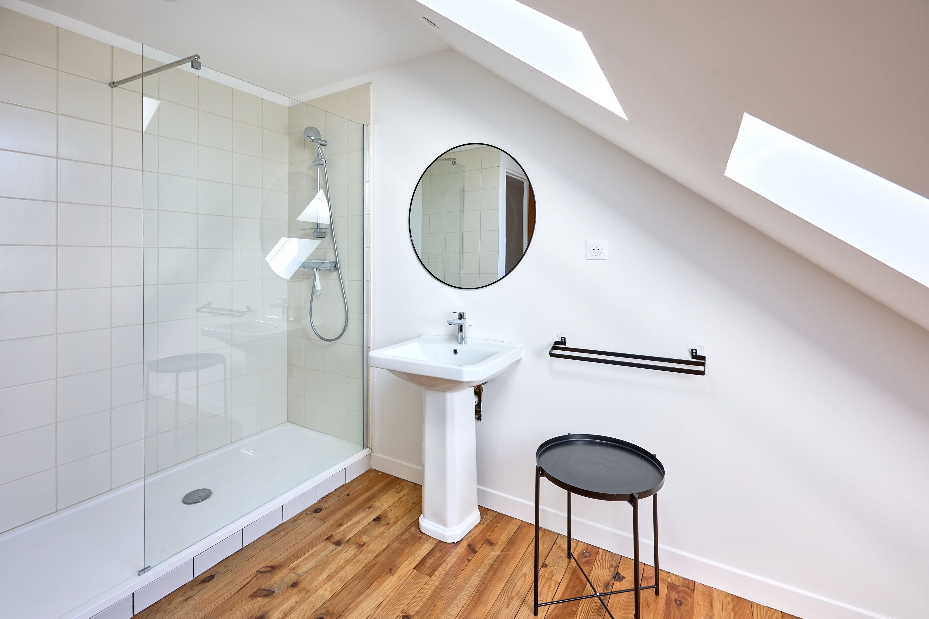 Un baño moderno con mampara de vidrio, un lavamanos pedestal blanco, un espejo redondo y un taburete negro pequeño.