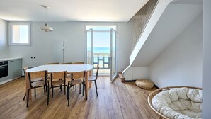 A modern kitchen with wooden flooring, a dining table with chairs, and a large window overlooking the sea.