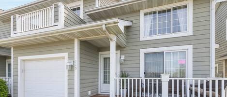 A two-story house with a covered front porch, a garage, and a small front yard with bushes.