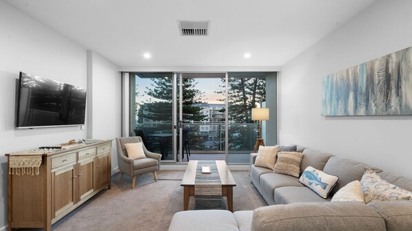 A modern living room with a flat-screen TV, wooden cabinet, sofa, and a view through glass doors to the outside. - Coast on Colley Terrace (Glenelg)