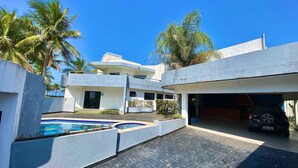 A modern multi-story building with a swimming pool, a garage, and palm trees. - Casa Alto Padrão Frente ao Mar (Peruibe)