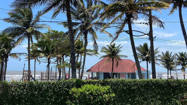 A coastal view with palm trees, a red-roofed building, and a beach in the background. - Casa Alto Padrão Frente ao Mar (Peruibe)