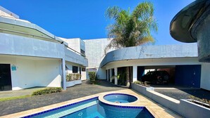 A swimming pool with a tiled edge, a vehicle in a garage, and a building with a curved roof. - Casa Alto Padrão Frente ao Mar (Peruibe)