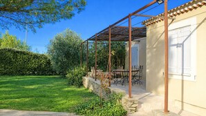 A covered outdoor seating area with a table and chairs, surrounded by a well-maintained garden and a building with a tiled roof.