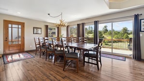 A dining area with a large wooden table and chairs, a chandelier, and a view of a green landscape through large windows.