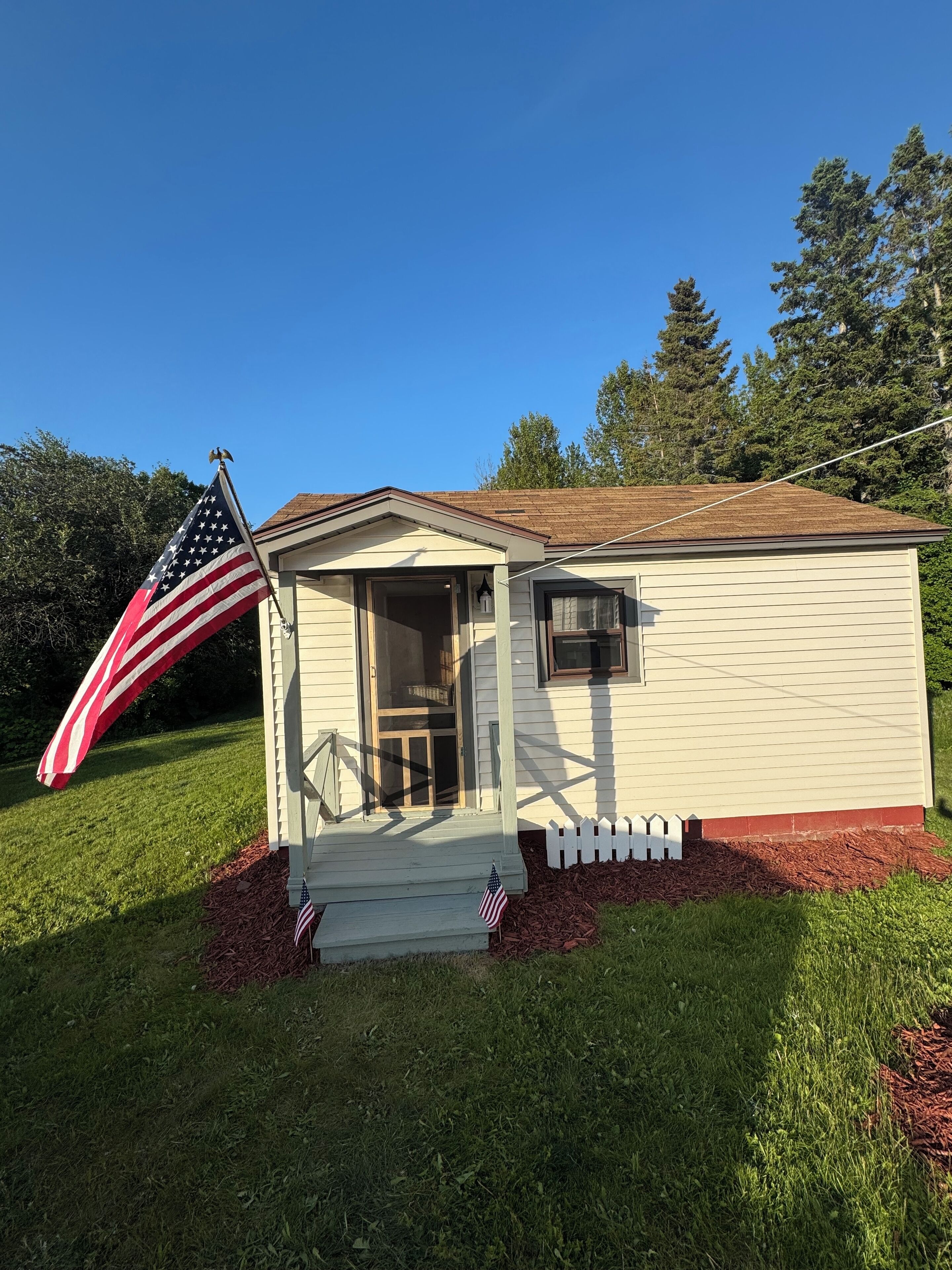 Una piccola casa a un piano con un portico e un albero con la bandiera americana.