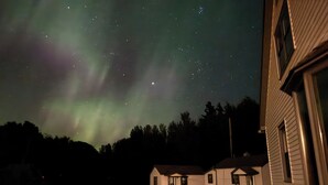 Talo, jossa on kuisti ja pieni varasto, ja sen yllä yötaivas, jossa näkyy tähtiä ja revontulia.