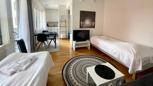 Habitación de hotel pequeña con cama, televisor, escritorio con silla y zona de cocina al fondo.