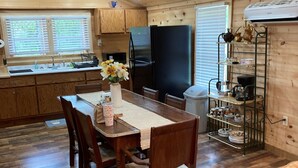 A wooden cabin interior with a dining table, chairs, a refrigerator, a sink, and a shelf with kitchenware.