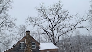 A wooden cabin with a stone chimney, a porch with a bench, and snow-covered surroundings.