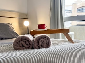 Una cama bien hecha con toallas enrolladas, una taza roja y una mesita de noche con lámpara.