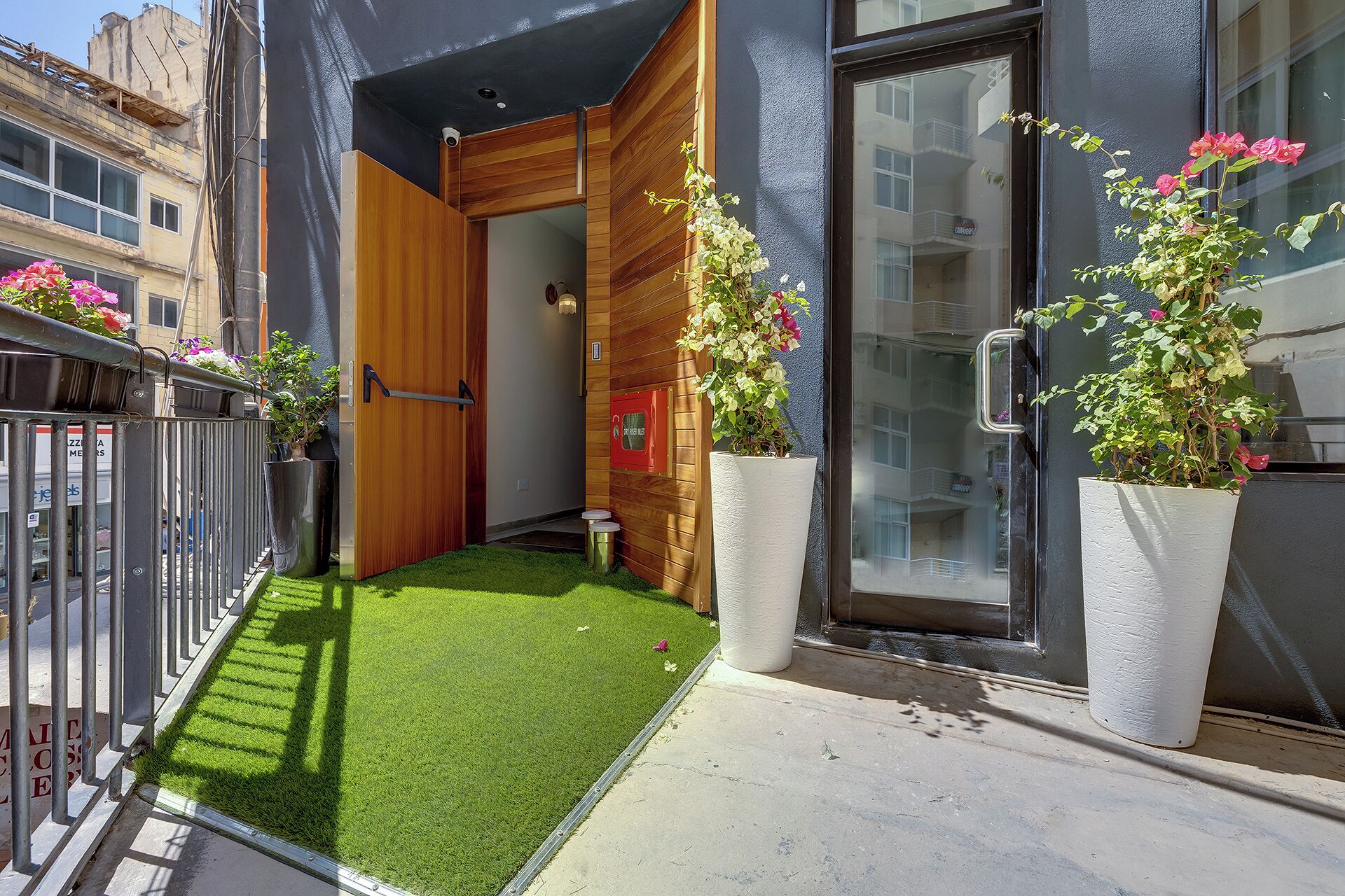 A modern building with a wooden door, a small balcony with artificial grass, and potted plants.