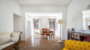 Een woonkamer met een bank, een gele fauteuil, een eettafel met stoelen, een vloerlamp en een boekenplank.
