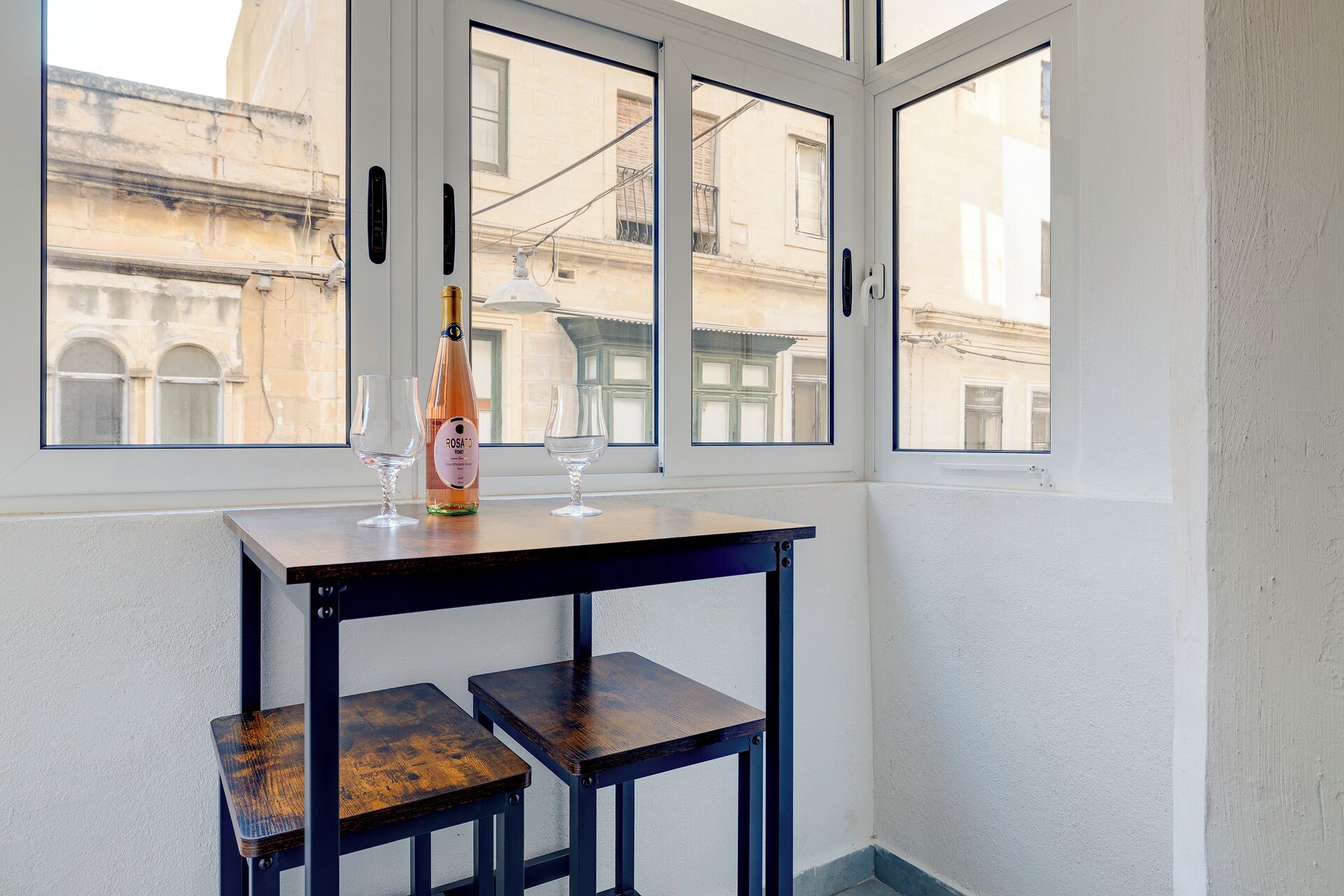 Une pièce d’angle avec une fenêtre donnant sur un immeuble, une table avec une bouteille et des verres, et deux tabourets en bois.