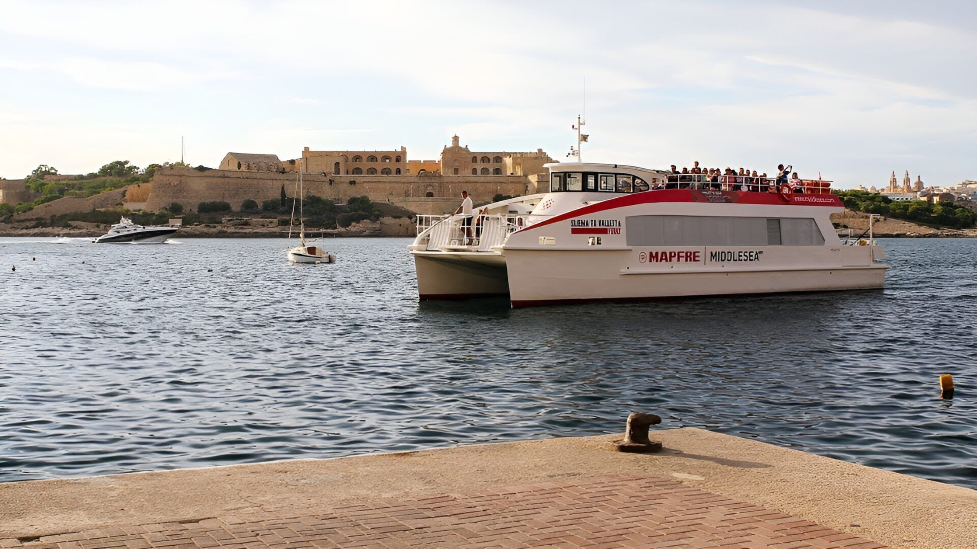 A ferry with 'MAPFRE MIDDLESEA' branding, docked at a pier with a historic building in the background.