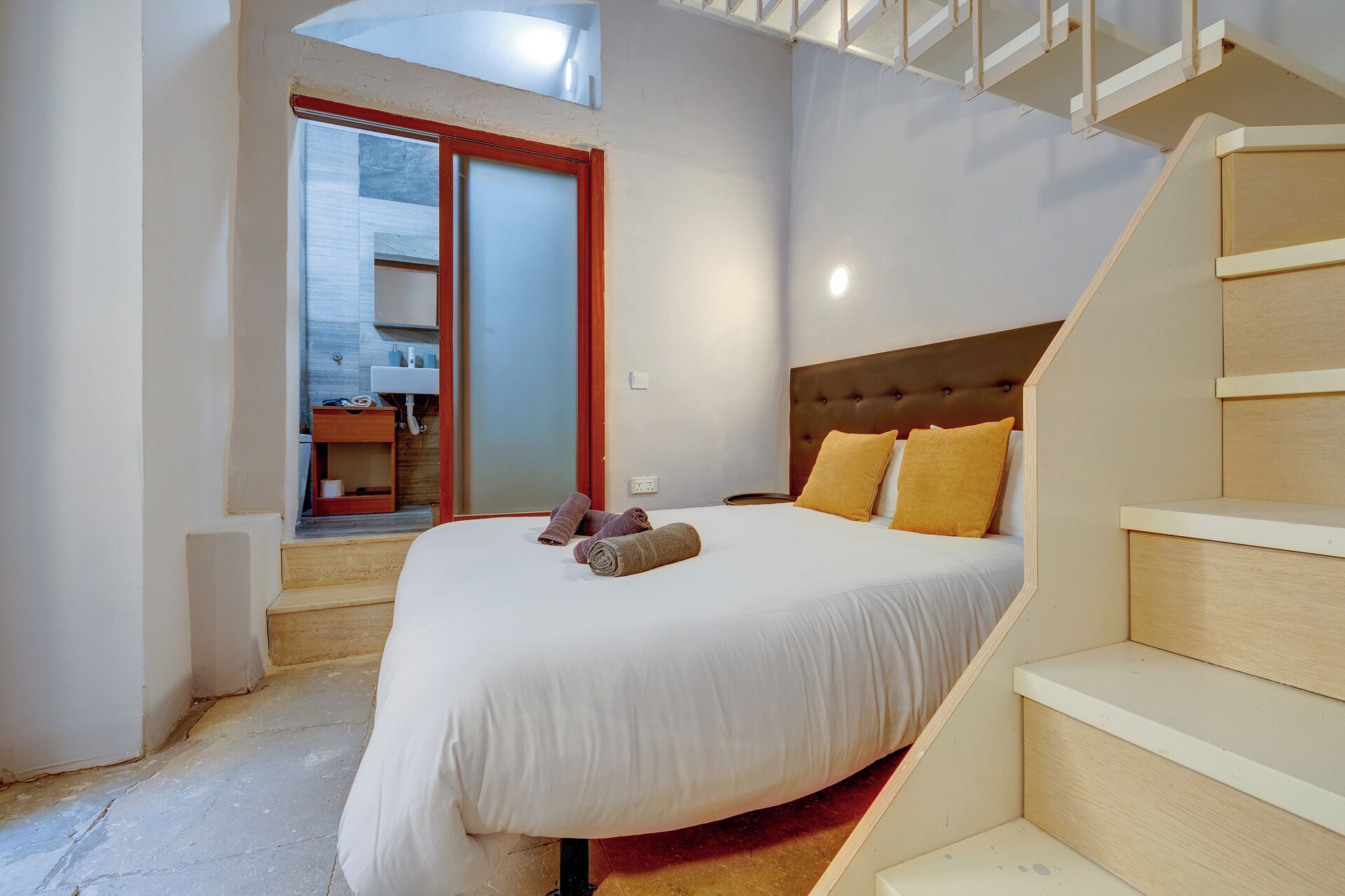 A bedroom with a bed, a wooden headboard, a staircase, and a bathroom visible through an open door.