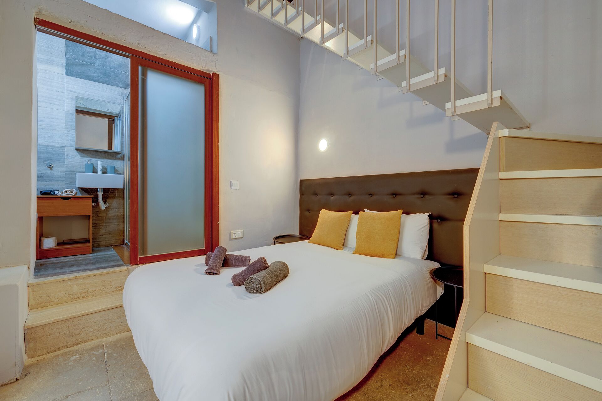 A bedroom with a bed, a nightstand, a wooden staircase, and a bathroom with a sink and mirror.