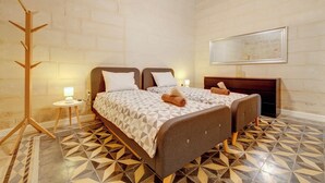 Una cama doble con colcha de estampado geométrico, un perchero de madera, una mesita de noche con lámpara y un espejo en la pared.