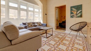 Amplia sala de estar con un conjunto de sofás, una mesa de centro y una silla. Hay ventanas grandes y una puerta que da a otra habitación.