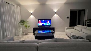 Una sala de estar moderna con un televisor de pantalla plana sobre un soporte negro, un sofá gris, una planta en maceta y dos luces fijadas a la pared.