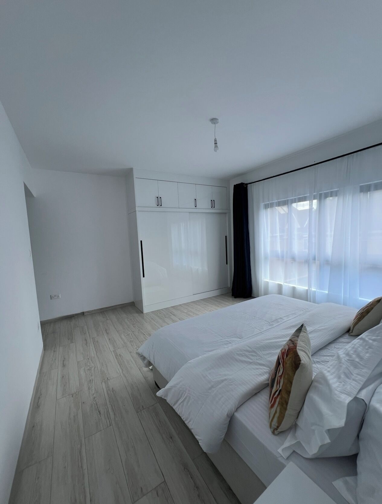 Ein Schlafzimmer mit einem großen Bett, einem weißen Kleiderschrank und einem Fenster mit transparenten Vorhängen.