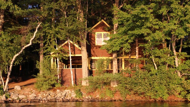 Une cabane dotée d’une grande fenêtre et d’une terrasse, entourée d’arbres et bordée par un plan d’eau.