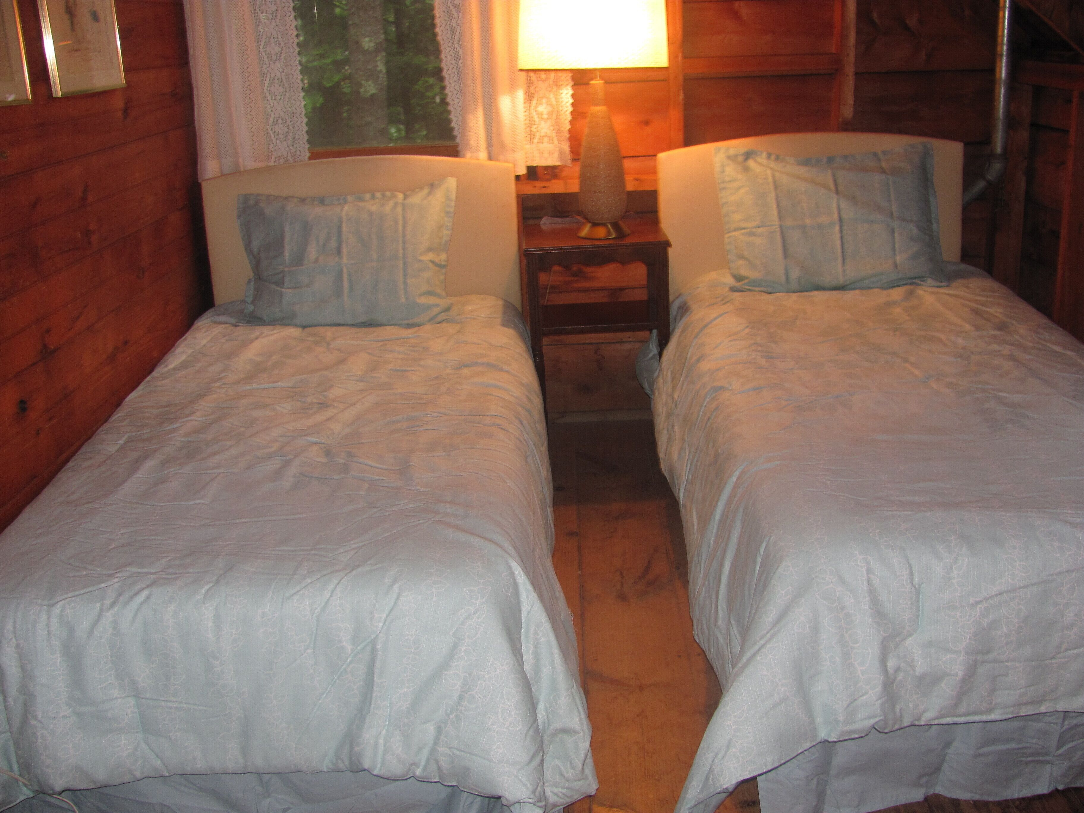 A room with two single beds, a nightstand with a lamp, and a window with lace curtains.