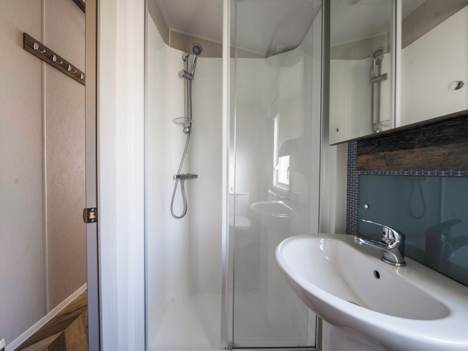 Een badkamer met een douchecabine van glas, een wit wastafelmeubel en een houten kastje met haken.