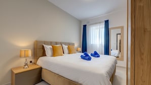A hotel room with a large bed, a bedside table with a lamp, a mirror, and a view of the outside through a window with curtains.