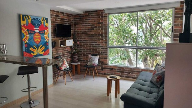 Una sala de estar moderna con pared de ladrillo, un ventanal grande, un sofá negro, una mesa de centro y un cuadro abstracto y colorido.