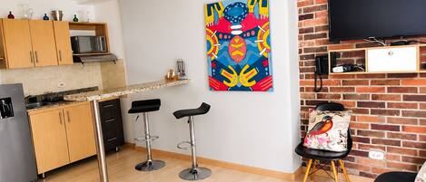 A modern kitchen with wooden cabinets, a microwave, and a countertop. There are two bar stools and a table with a remote control. A colorful abstract painting is on the wall, and a television is mounted on a brick wall.