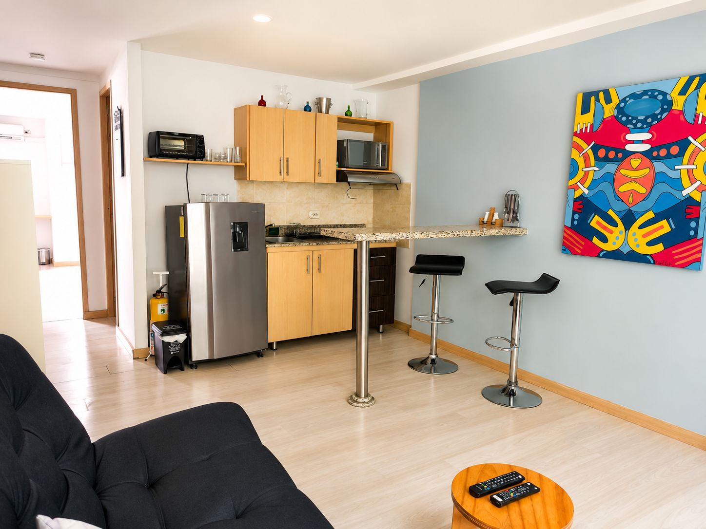A modern kitchen with a refrigerator, microwave, and wooden cabinets. There are two bar stools and a painting on the wall.