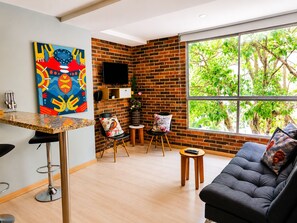 A modern living space with a brick wall, a large window with a view of trees, a black sofa, a colorful abstract painting, and a small wooden table. - Medellin, Bring your toothbrush, in Laureles (Medellín)