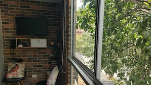 Una habitación con pared de ladrillo, un televisor de pantalla plana montado sobre un mueble de madera, una ventana con vistas a zonas verdes y dos sillas con cojines estampados.