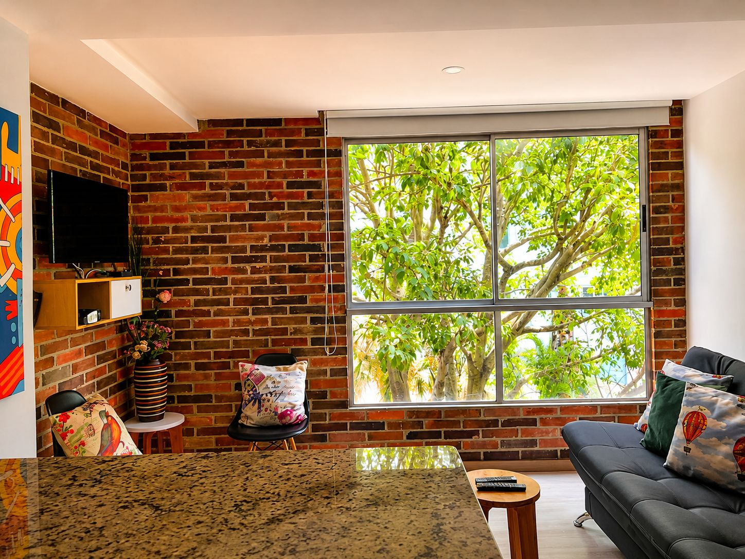 A living room with a brick wall, a large window with a view of trees, a sofa, and a TV mounted on the wall.