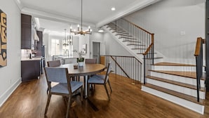 A spacious dining area with a wooden table and chairs, a modern kitchen in the background, and a staircase with wooden steps and black railings.