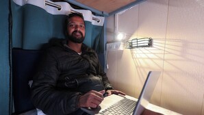 A person using a laptop in a room with a bed, a curtain, and a wall-mounted light fixture. - Laxmi Dormitory | 2 mins walk from Prem Mandir (Vrindavan)