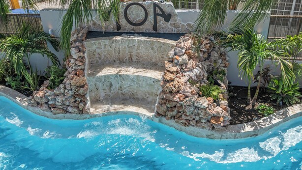 A pool with a stone wall and palm trees. - NÔA curacao (Willemstad)
